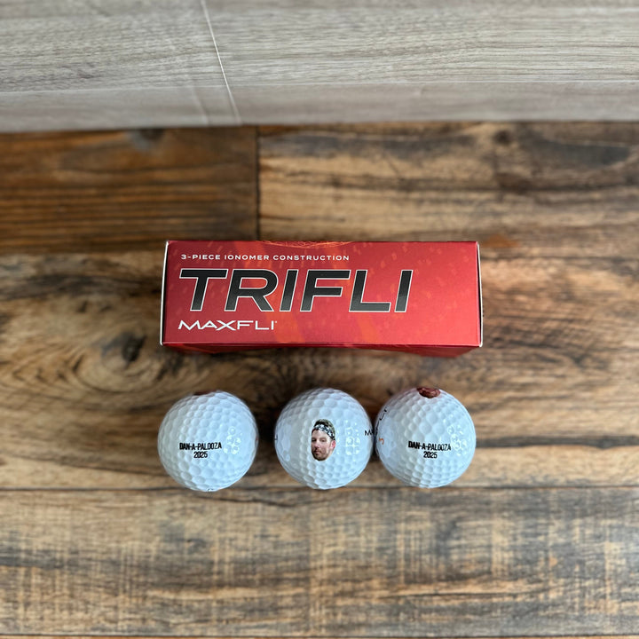 Three golf balls in front of a red Maxfli Trifli box on a wooden surface