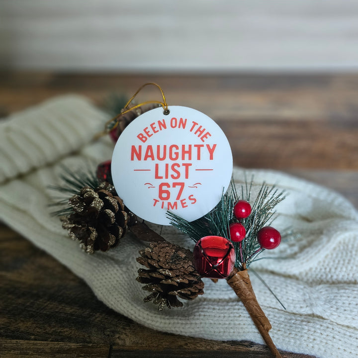 Decorative Christmas ornament with a tag reading 'Been on the Naughty List 67 Times' on a textured surface.