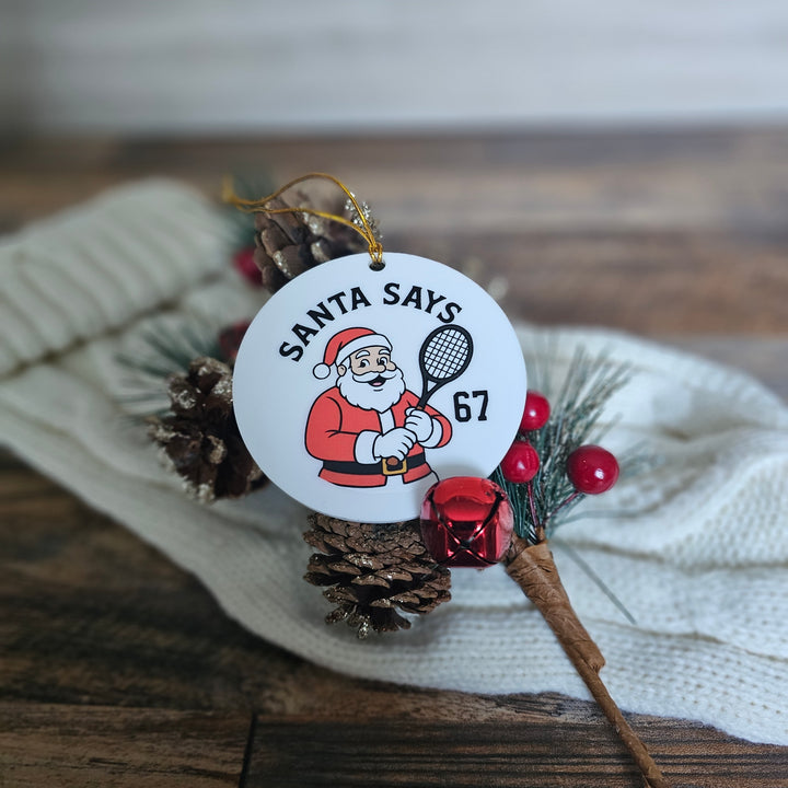 Christmas ornament with Santa Claus holding a tennis racket on a wooden surface with pine cones and berries.