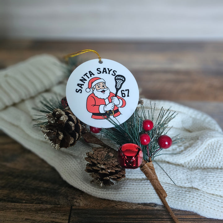 Christmas ornament with Santa Claus design and text 'Santa Says 67' on a wooden surface.