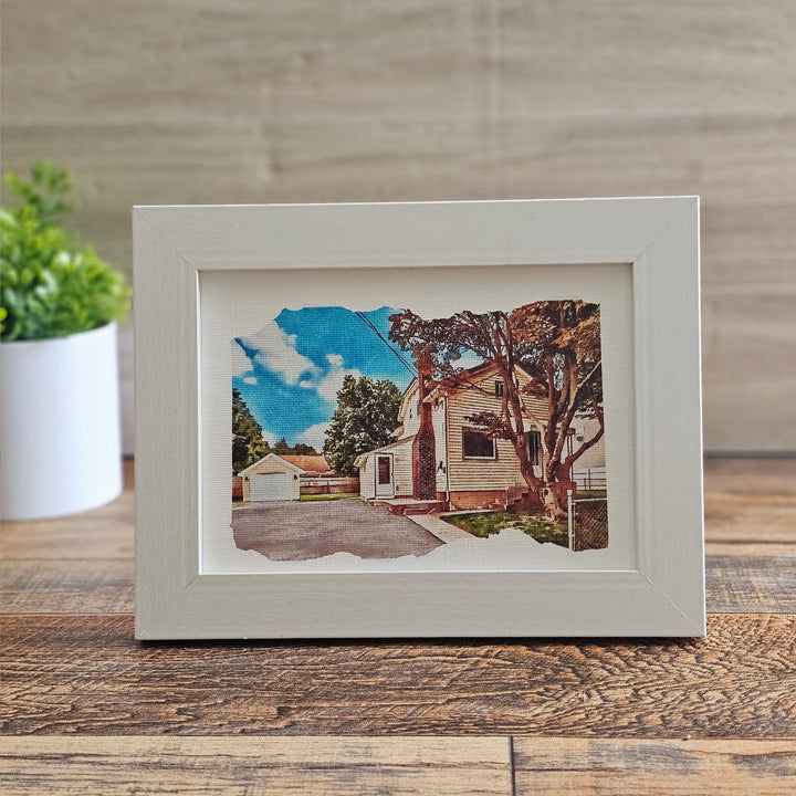 Framed artwork of a house on a wooden surface with a plant in the background