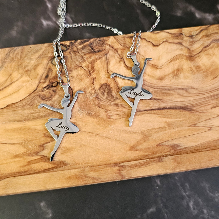 Two silver ballerina pendants with engraved names on a wooden surface