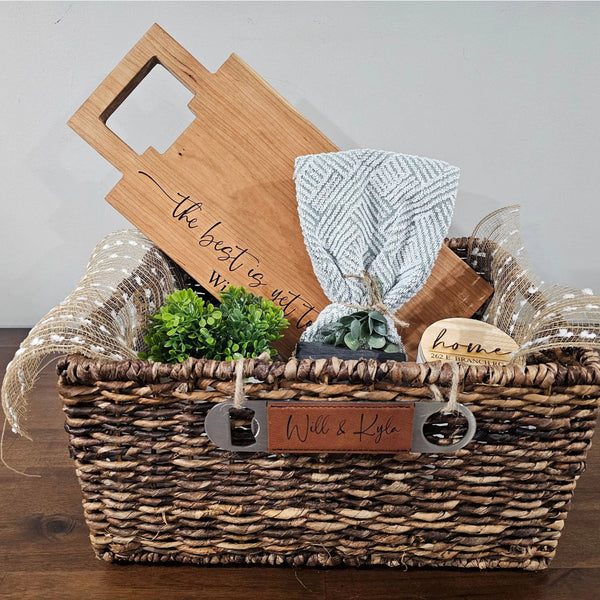 Wicker gift basket with wooden cutting board, glass, and other items on a wooden surface.