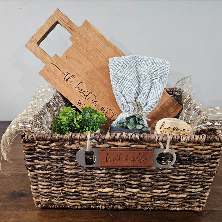 Wicker gift basket with wooden cutting board, glass, and other items on a wooden surface.