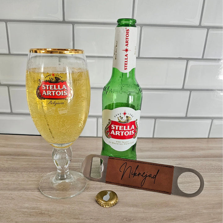Stella Artois bottle, glass, and opener on a wooden surface with a tiled wall background