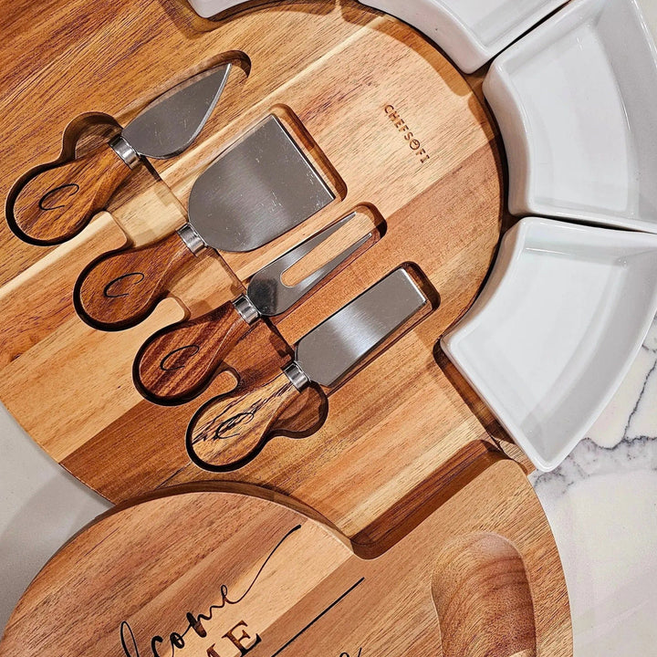 Wooden cheese board set with engraved text and four cheese knives on a marble surface.