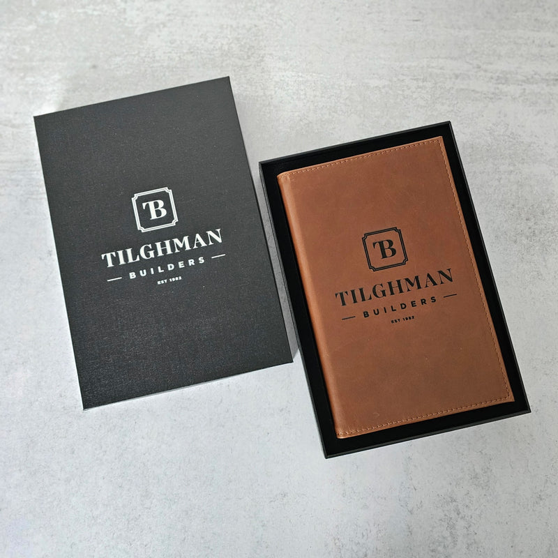Brown leather notebook with 'Tilghman Builders' branding on a gray surface